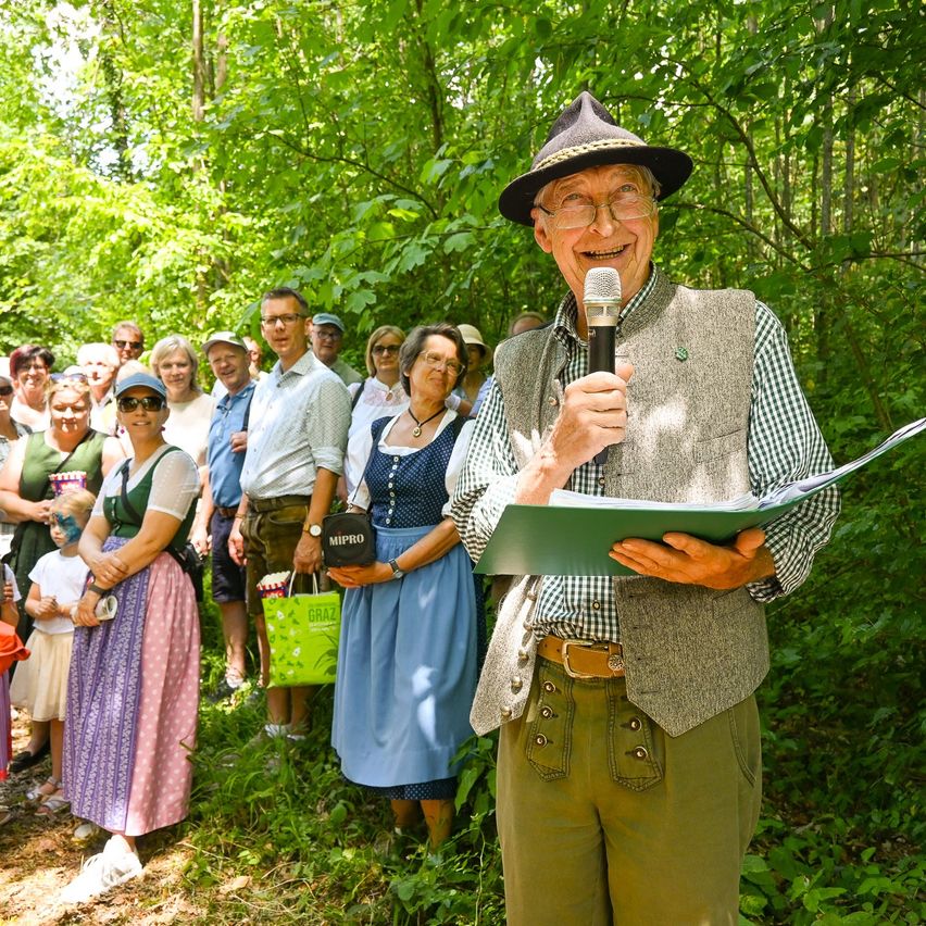 Ein älterer Mann in traditioneller bayerischer Kleidung spricht in einen Mikrofon in einem Wald, umgeben von einer Gruppe von Menschen in traditioneller Kleidung.