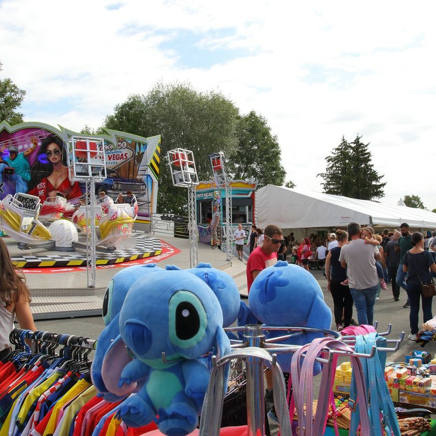 Ein geschäftiger Jahrmarkt mit einer großen Fahrt, die eine Frau in einem roten Kleid zeigt, umgeben von Menschen, die einkaufen und die Attraktionen genießen.