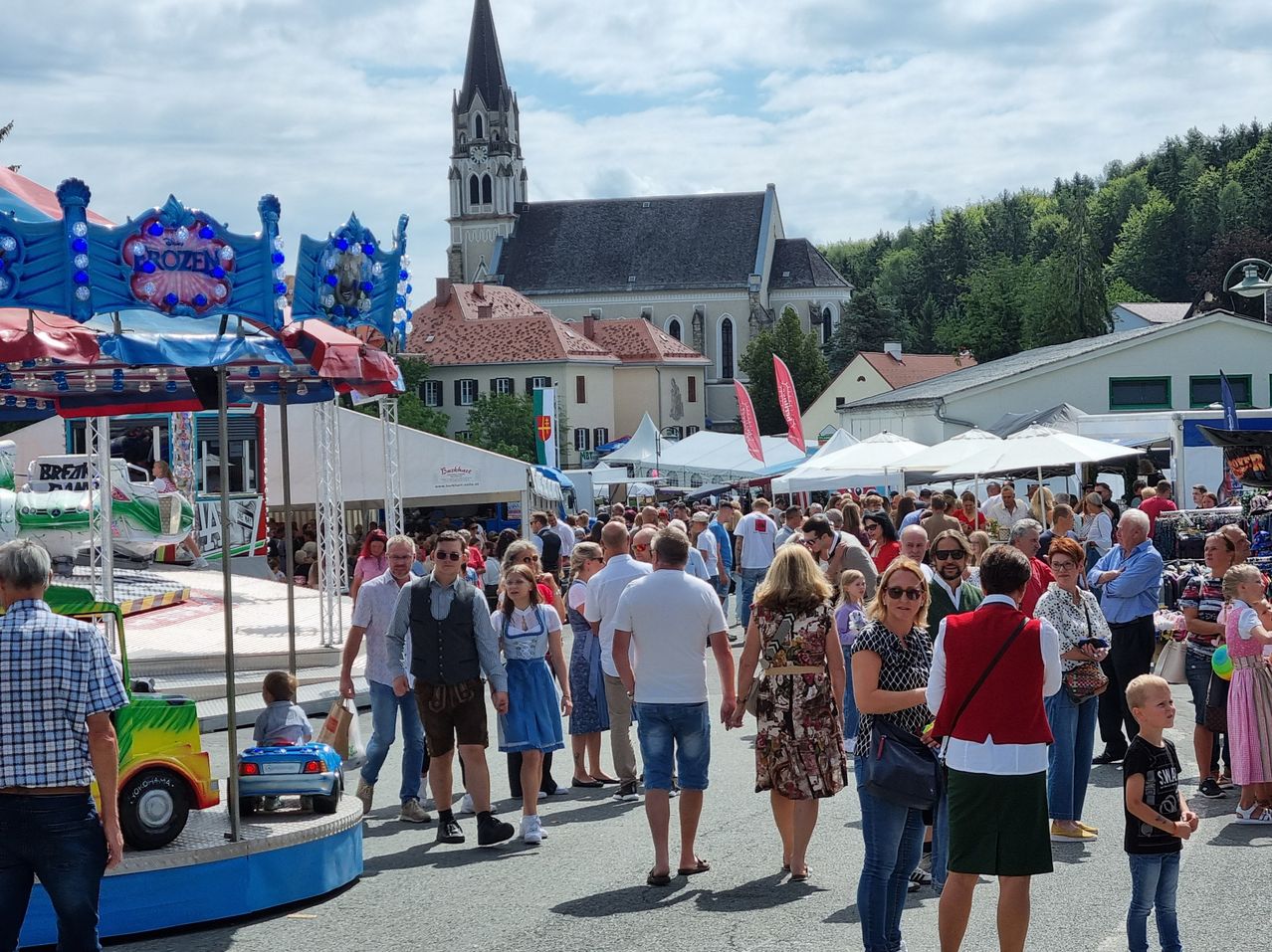 Ein geschäftiges Freiluft-Event mit einer Menge Menschen, die an einem Karussell, weißen Zelten und einer Kirche im Hintergrund vorbeigeht.