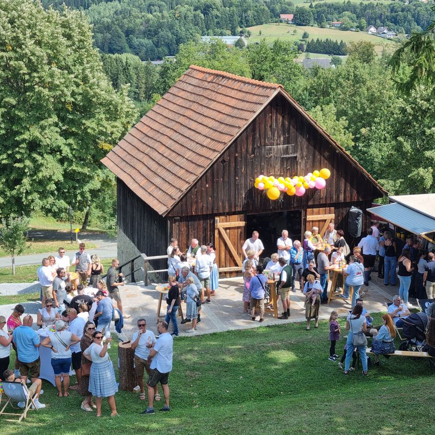 Eine Gruppe von Menschen versammelt sich vor einem rustikalen Bauernhaus, mit bunten Luftballons, die vom Dach hängen. Sie stehen um Tische herum und unterhalten sich.