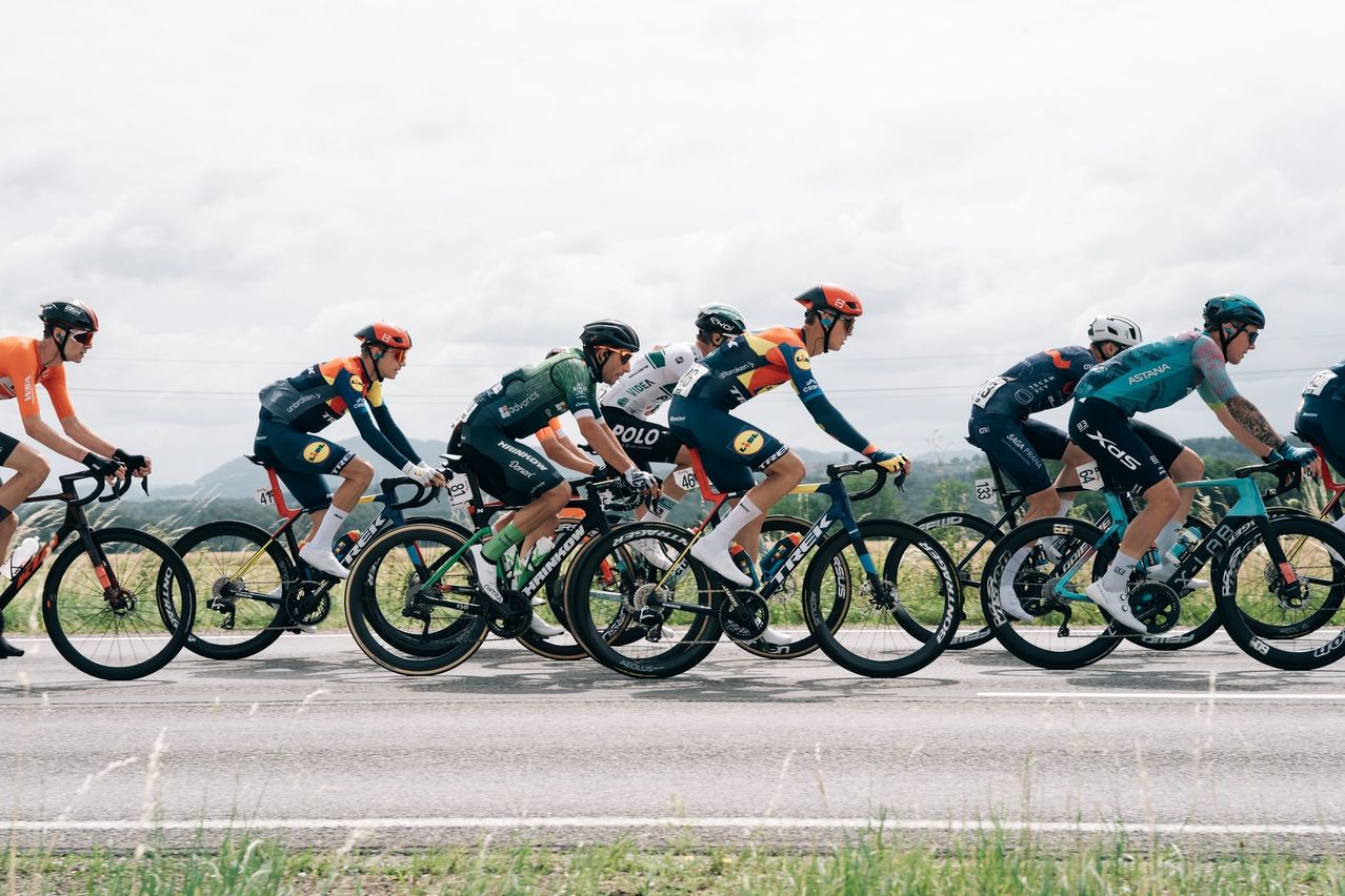 Cyclists in team jerseys compete in a race, riding on a paved road with grass on the sides. Helmets and cycling gear are visible.