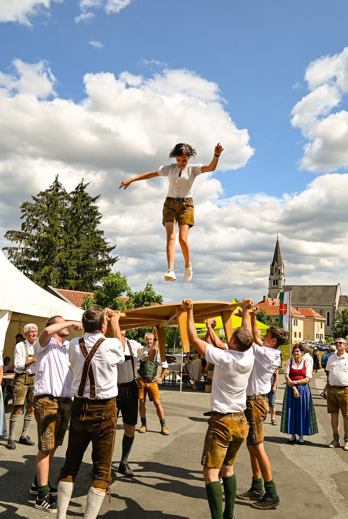 A woman jumps over a table held by several men in traditional clothing, outside near a church.