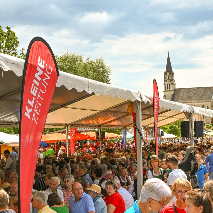 A large crowd gathers under white tents, with red banners reading 'Kleine Zeltung' and a church visible in the background.