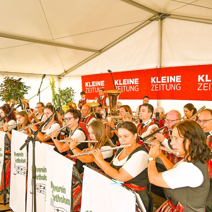 A group of musicians dressed in traditional attire are performing on stage with wind instruments under a tent. In the background, a red banner reads 'Kleine Zeitung'.