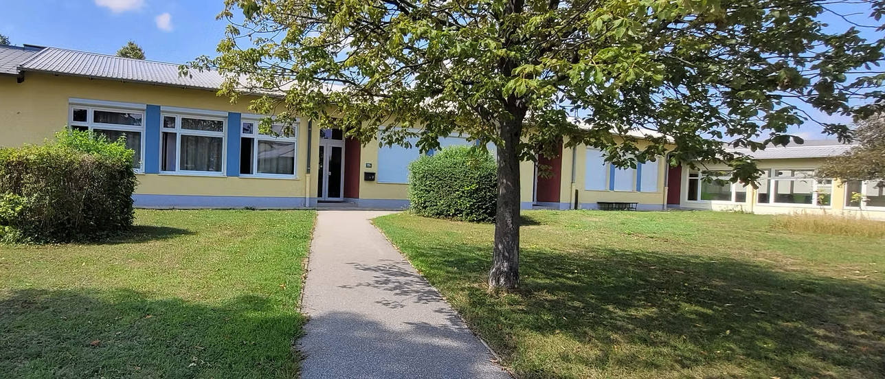 Ein gelbes Gebäude mit blauen Fensterläden und einem Gehweg, der zu seinem Eingang führt. Der Eingang wird von zwei kleinen Büschen flankiert. Ein Baum steht rechts vom Weg.