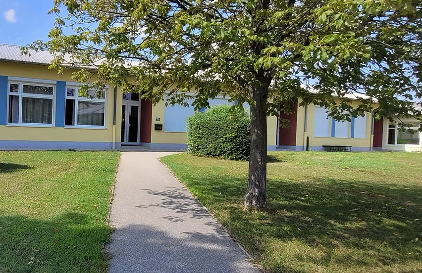 Ein gelbes Gebäude mit blauen Fensterläden und einem Gehweg, der zu seinem Eingang führt. Der Eingang wird von zwei kleinen Büschen flankiert. Ein Baum steht rechts vom Weg.