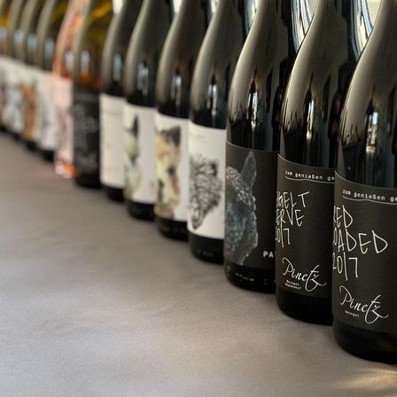 Eine Reihe von Weinflaschen mit schwarzen Etiketten, auf einem Tisch ausgestellt. Die Etiketten enthalten Illustrationen und Text. Die Flaschen sind in einer Reihe angeordnet.