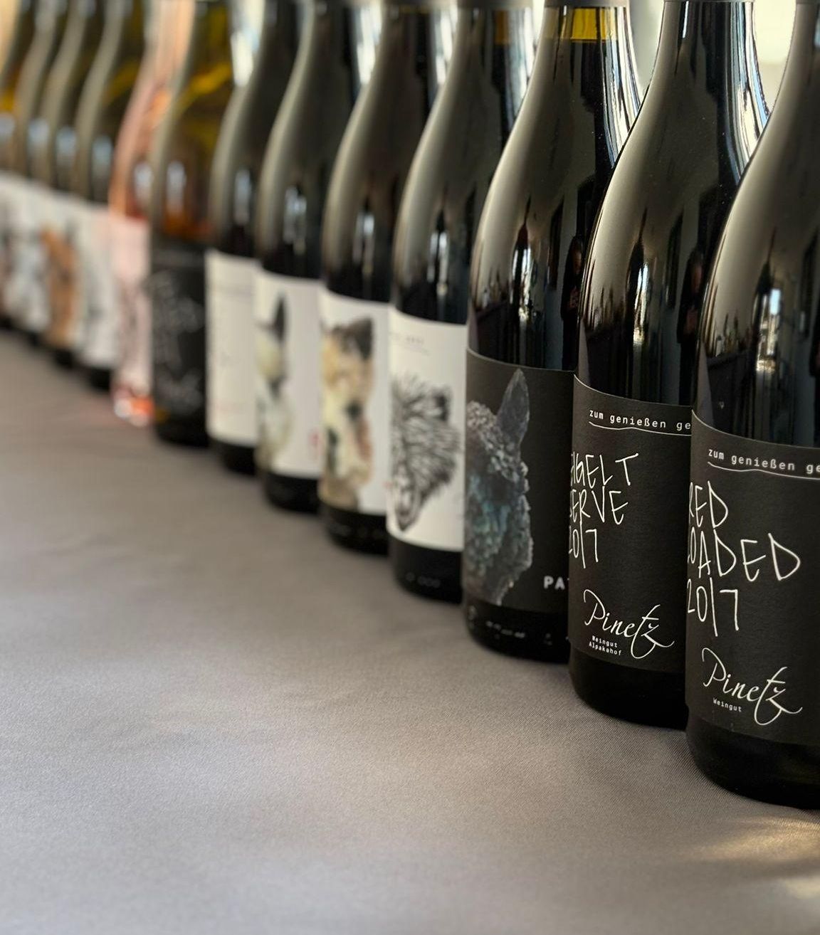 Eine Reihe von Weinflaschen mit schwarzen Etiketten, auf einem Tisch ausgestellt. Die Etiketten enthalten Illustrationen und Text. Die Flaschen sind in einer Reihe angeordnet.