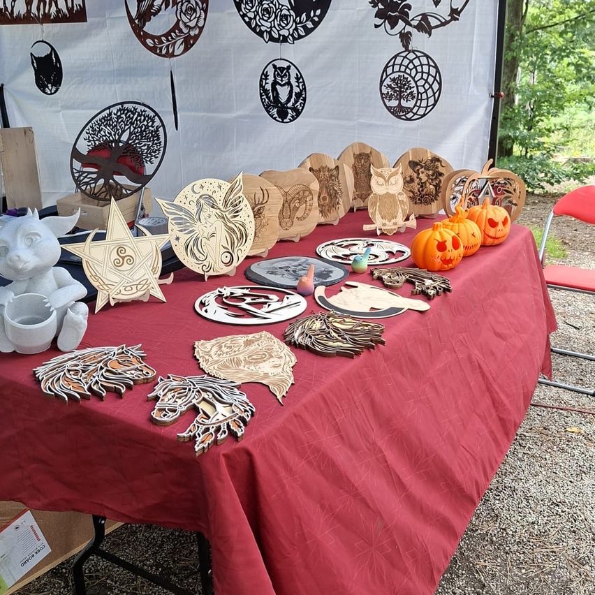 Ein Tisch mit roter Tischdecke zeigt verschiedene Holzschnitzereien, darunter Tiere, Sterne und Kürbisse. Dahinter befindet sich ein weißer Hintergrund mit hängenden Dekorationen. Die Umgebung ist im Freien mit Bäumen im Hintergrund.