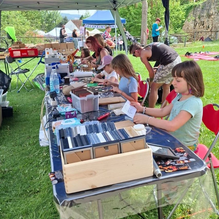 Eine Gruppe von Kindern und Erwachsenen versammelt sich an einem Festivaltisch. Sie sind mit einer Bastelaktivität beschäftigt, umgeben von verschiedenen Materialien. Ein Zelt befindet sich in der Nähe, und im Hintergrund sind Menschen und Zelte zu sehen.