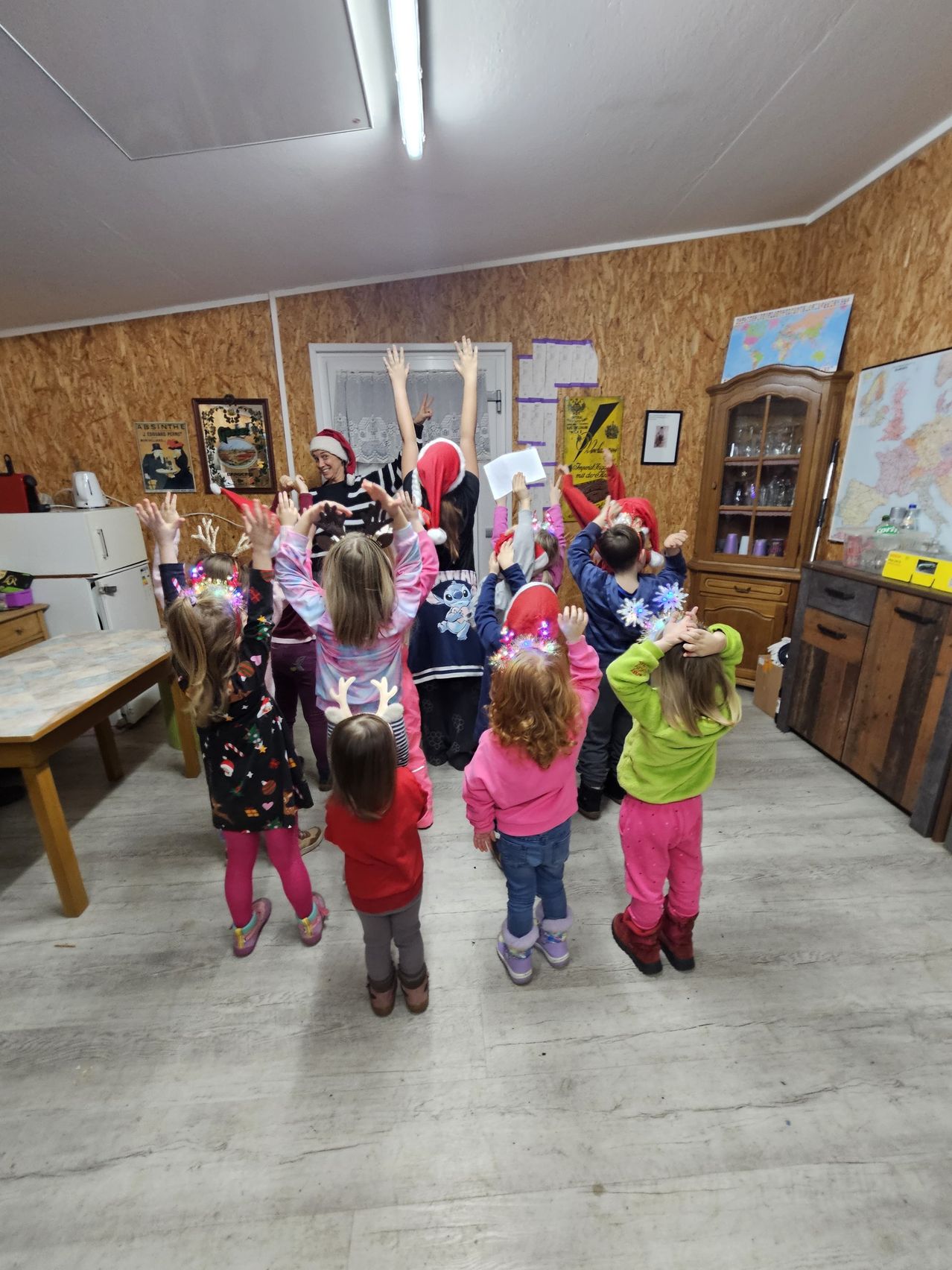 Eine Gruppe von Kindern in einem Klassenzimmer mit Holzwänden und einem Holzboden. Sie tragen Weihnachtsmützen und haben ihre Hände erhoben. Eine Frau in einer Weihnachtsmütze steht vor ihnen.