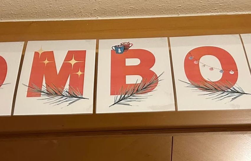 Drei Buchstaben, die das Wort M B O ergeben, stehen auf einem Holzregal. Der Buchstabe M hat ein Tannenzapfendesign, B hat ein Kaffeetassendesign und O hat ein Lichterkettendesign.