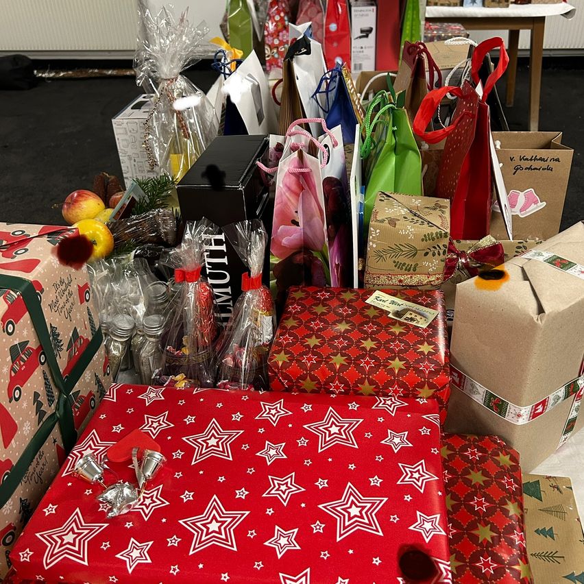 Verschiedene verpackte Geschenke auf einem Tisch, darunter rot-gestirntes Geschenkpapier, durchsichtige Plastiktüten und braune Pappkartons. Einige haben Bänder und Etiketten. Der Tisch ist mit vielen verschiedenen verpackten Geschenken überladen.