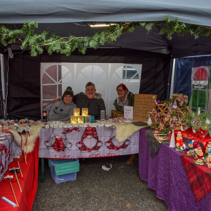 Three people sit under a canopy, smiling, with Christmas decorations around them. A table in front has items on it, including candles and trinkets. A Christmas tree is on the left, and a wooden signboard is on the right.