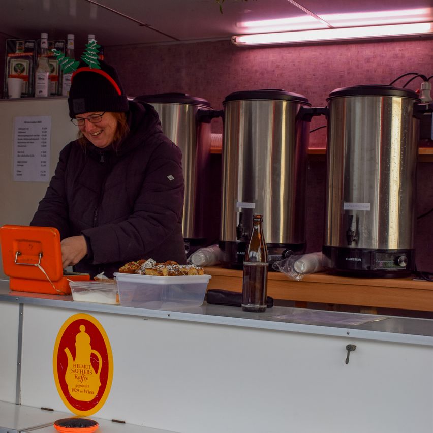 A woman in a black jacket and glasses is standing behind a counter, smiling. Three stainless steel coffee urns are behind her, and a plastic container with food is on the counter. There is a sign with a teapot logo and text on the counter.
