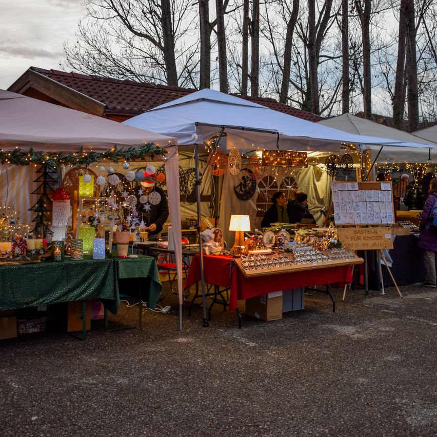 A Christmas market setup with multiple stalls under white canopies. Various holiday items, including ornaments, are displayed. People are browsing and shopping at the stalls.