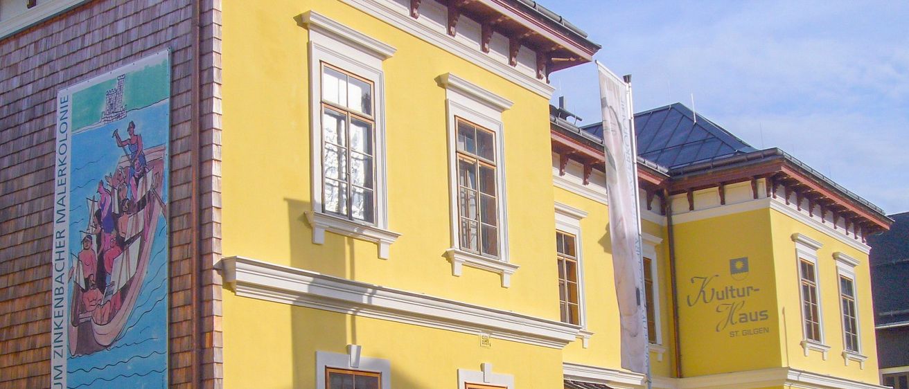 Ein leuchtend gelbes Gebäude mit weißen Umrandungen und Glasfenstern. Links eine Wandmalerei, rechts eine Flagge. Der Text lautet Kultur-Kastel St. Gilgen.