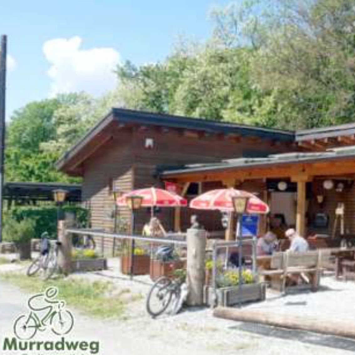 Ein Outdoor-Restaurant mit hölzerne Außenwand und roten Sonnenschirmen, umgeben von einigen Holztischen und Stühlen, mit sitzenden Menschen und einem geparkten Fahrrad.