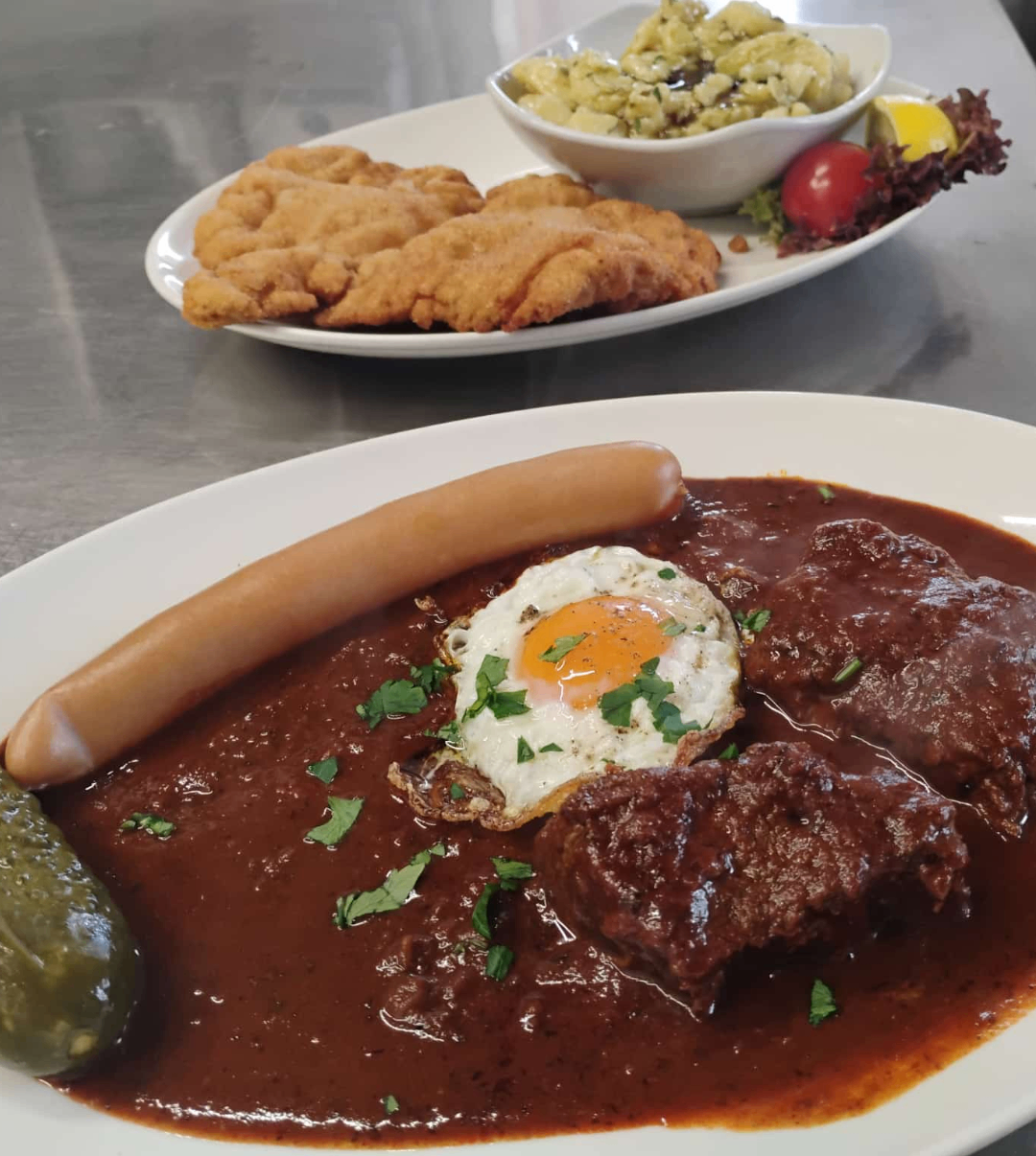 Zwei Teller mit Essen auf einem Tisch. Ein Teller hat ein Fleischgericht mit einem Spiegelei und einer Wurst. Ein anderer Teller hat ein gebratenes Brot mit Käse.
