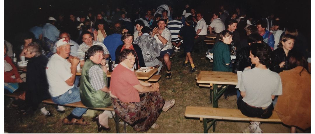 Eine Zusammenkunft von Menschen in einer ungezwungenen Umgebung, die bei Nacht auf einem Rasenfeld um Tische herum sitzen. Flaschen und Teller stehen auf den Tischen.