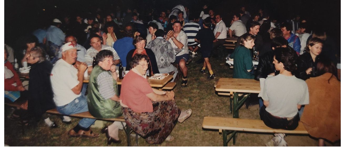 Eine Zusammenkunft von Menschen in einer ungezwungenen Umgebung, die bei Nacht auf einem Rasenfeld um Tische herum sitzen. Flaschen und Teller stehen auf den Tischen.