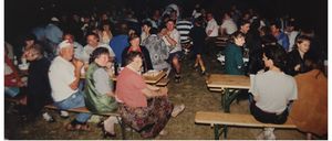 Eine Zusammenkunft von Menschen in einer ungezwungenen Umgebung, die bei Nacht auf einem Rasenfeld um Tische herum sitzen. Flaschen und Teller stehen auf den Tischen.