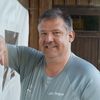 A man with a smile stands outside, wearing a gray shirt with white text, next to a wooden door.