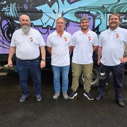 Vier Männer in weißen Poloshirts mit Logos stehen vor einem bunt besprühten Lastwagen.