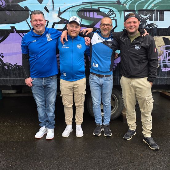 Vier Männer stehen vor einem bunten Graffiti-Truck in Sportjacken und Sneakers.