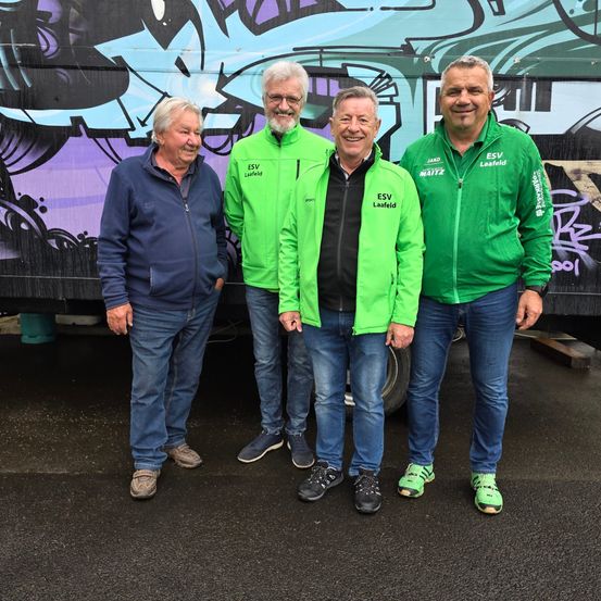 Vier Männer stehen vor einem Bus mit Graffiti an der Seite. Sie tragen alle Jacken und Jeans. Der Mann auf der linken Seite trägt eine blaue Jacke, während die anderen grüne Jacken tragen.