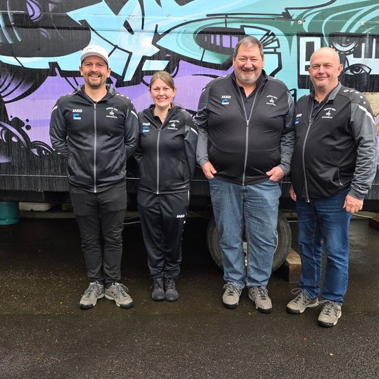 Vier Personen posieren für ein Foto vor einer mit Graffiti bemalten Wand. Sie tragen alle schwarze Jacken mit Text und Logos.