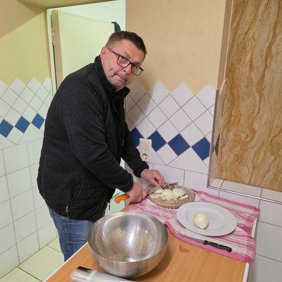 Ein Mann bereitet Essen in einer Küche zu. Er trägt eine Brille und eine Jacke. Er schneidet Käse auf einem Teller.