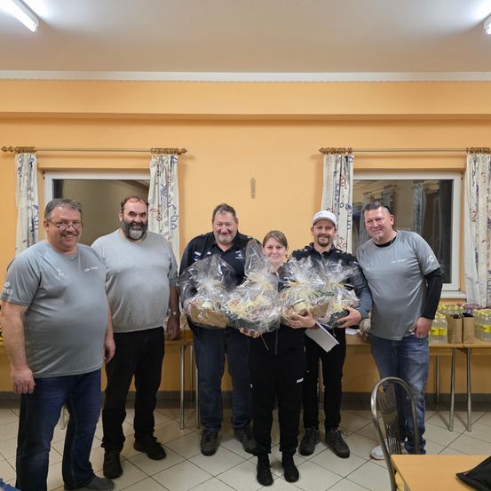 Fünf erwachsene Männer und eine Frau posieren für ein Foto in einem Raum mit zwei Tischen und halten Lebensmittelpakete in der Hand.