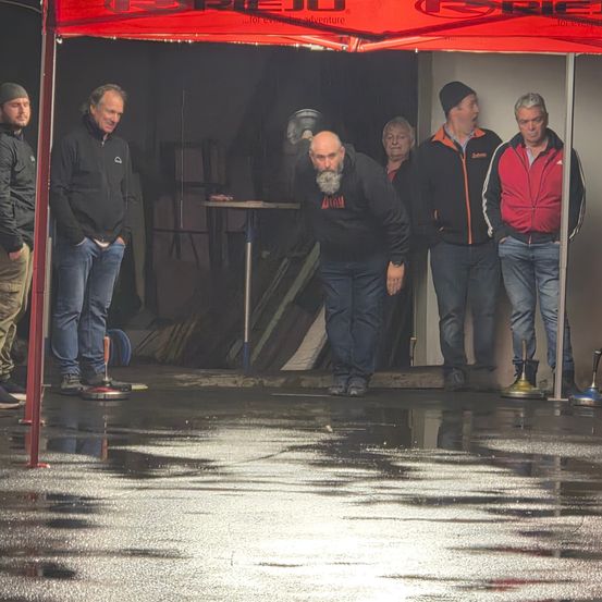 Sechs Männer in Jacken und Jeans stehen in einem Zelt unter einem roten Baldachin. Ein Mann steht vorne, mit den Händen in den Taschen. Der Boden ist nass.