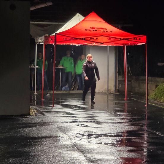 Eine Person in einem roten Zelt geht auf einer nassen Straße, wobei Personen in grünen Jacken hinter dem Zelt stehen.