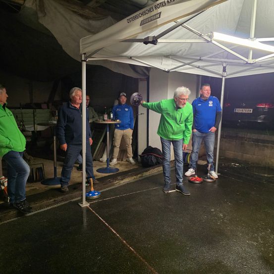 Eine Gruppe von Männern steht in einem Zelt auf einem nassen Betonboden. Der Mann in der Mitte wirft einen Ball in Richtung des Ziels, während die anderen zusehen.