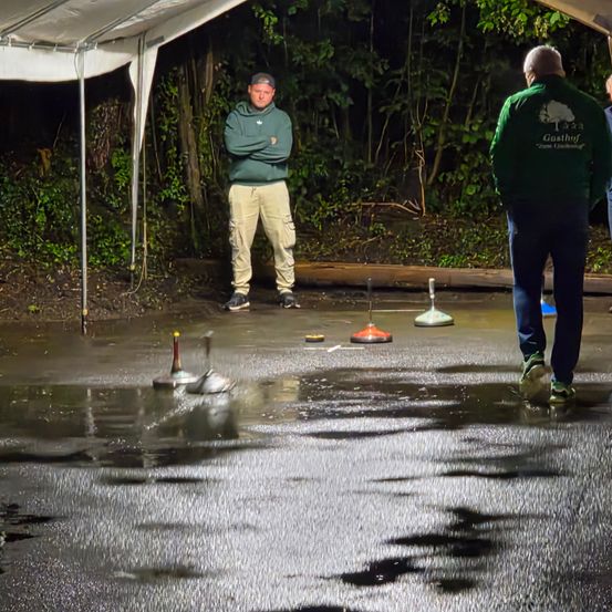 Zwei Männer stehen auf einem nassen Boden, vielleicht in einem Park. Einer von ihnen schaut den anderen Mann an, während der andere auf den Boden schaut.