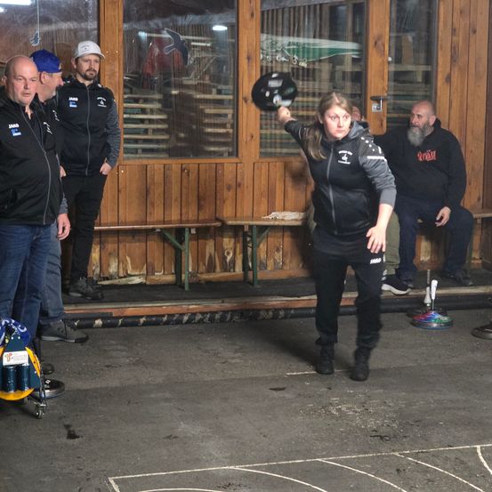 Eine Frau wirft einen Bowlingball auf die Pins in einer Bowlingbahn. Sie wird von mehreren Männern umgeben, einige stehen und einige sitzen auf einer Bank.