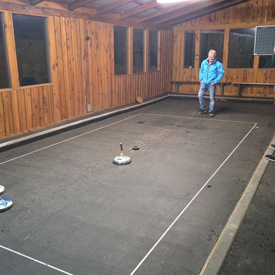 Ein Mann in einer blauen Jacke steht auf einem Platz mit einer Scheibe, drinnen, an einer Holzwand mit Fenstern und Bänken.