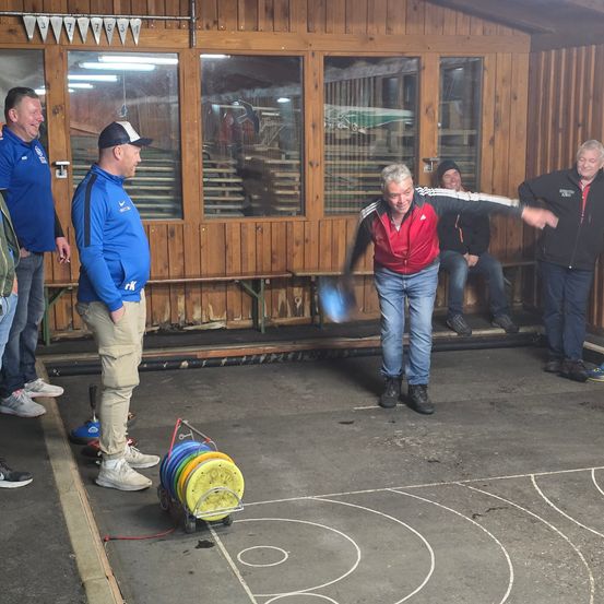 Ein älterer Mann in einem roten Jackett wirft einen Scheiben im Spiel. Vier andere Männer stehen und beobachten.