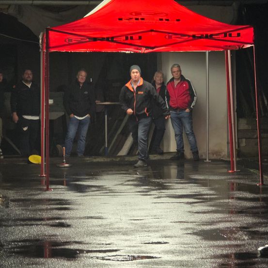 Eine Gruppe von Männern in Jacken und Jeans steht unter einem roten Zelt, einer wirft einen gelben Frisbee.