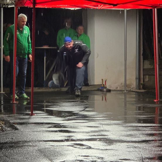 Eine Gruppe von Männern in grünen Jacken und blauen Mützen spielt unter einem roten Zelt. Der Boden ist nass, und der Mann in der Mitte wirft etwas.