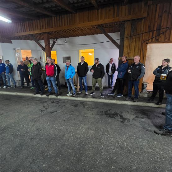 Eine Gruppe von Menschen steht in einer Reihe auf einem Betonsteig mit hölzernen Wänden im Hintergrund.