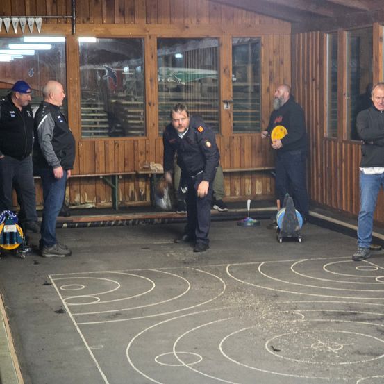 Fünf Männer stehen in einem Raum mit einer Holzwand. Ein Mann hält ein gelbes Frisbee, während ein anderer Mann auf dem Boden mit einem Spiralmuster geht.