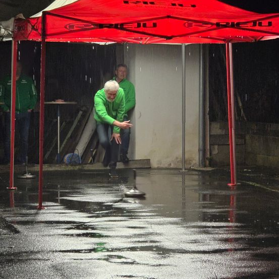 Ein Mann wirft einen Frisbee auf einer nassen Straße unter einem roten Zelt. Zwei Männer stehen hinter ihm.