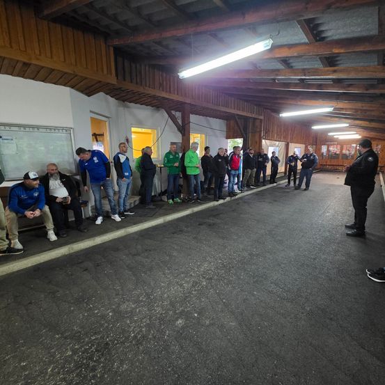 Sporthalle mit mehreren Personen, die entlang der Seitenlinien stehen, ein Mann spricht vorne