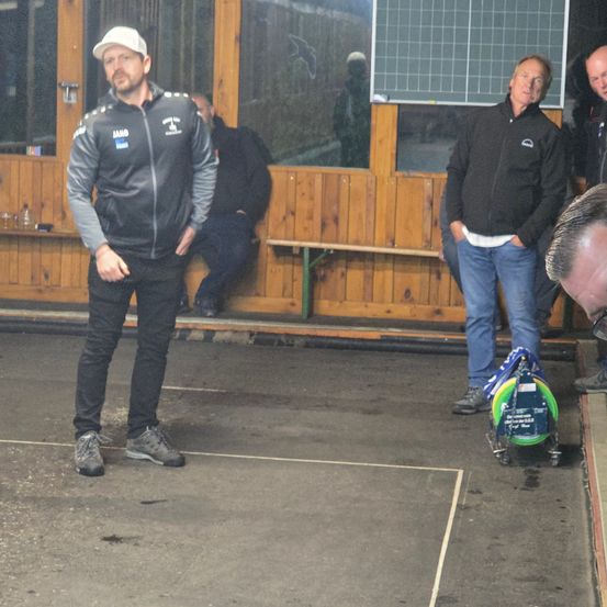 Ein Mann in einer schwarzen Jacke mit Logo steht auf einem Betonboden mit weißer Linie. Er schaut einen Mann an, der hinter ihm steht und ebenfalls eine schwarze Jacke und Jeans trägt. Der Mann hinter ihm hält ein grünes Objekt. Rechts schaut ein Mann auf den Boden.