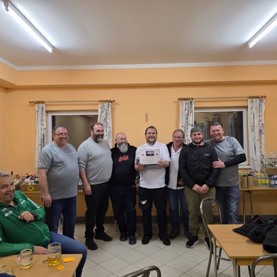 Eine Gruppe von Männern posiert für ein Foto mit einem Mann, der ein Zertifikat in einer Hand hält, in einem Raum mit Stühlen, Tischen und Flaschen Bier.