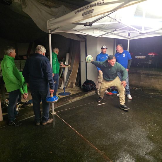 Fünf Männer in verschiedenfarbigen Jacken spielen Bocce auf einem nassen Bürgersteig. Ein Mann wirft den Pallino, während die anderen zuschauen.