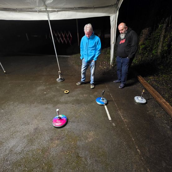 Zwei Männer stehen bei Nacht unter einem Zelt auf einer nassen Straße. Einer von ihnen hält einen Stock und sie spielen ein Spiel mit bunten Scheiben auf dem Boden.